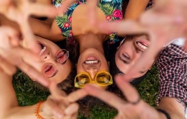 top view from above on colorful stylish happy young company of friends lying on grass in park, man and women having fun together, summer hipster fashion style, traveling smiling and showing peace sign